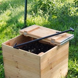 Un aérateur composteur de type flèche aérateur noir posé sur le couvercle en bois partiellement ouvert d'un bac à compost rempli de compost organique dans un jardin.